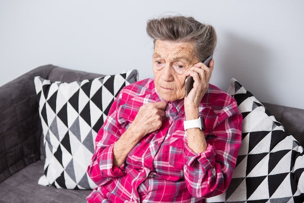 Una anciana anciana abuela con canas se sienta en su casa en el sofá usando el teléfono de mano