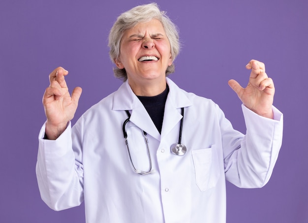 Anciana ansiosa en uniforme médico con estetoscopio se encuentra con los dedos cruzados