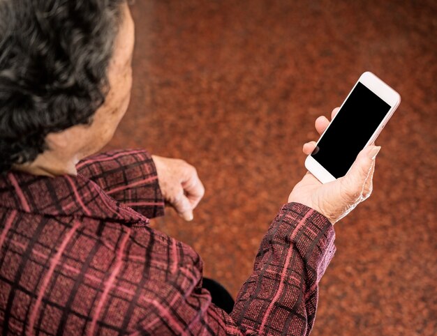 Anciana asiática sentada y mirando a través de algo en el teléfono inteligente moderno, haciendo conexión con otros en el hogar, tecnología viva, de cerca