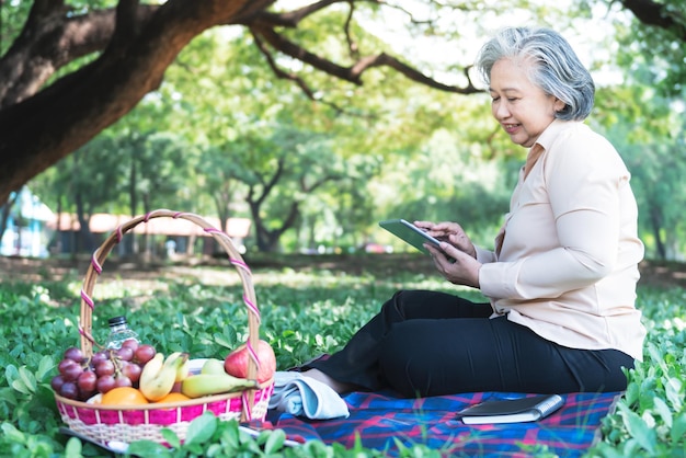 Anciana asiática sentada y relajada usando una tableta en el césped verde en el jardín con feliz