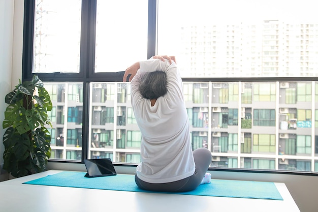 Foto una anciana asiática se sienta a hacer ejercicio en casa. hace posturas de yoga según un profesor de fitness en línea a través de una videollamada a través de una tableta. distanciamiento social que mantiene la salud de los ancianos.