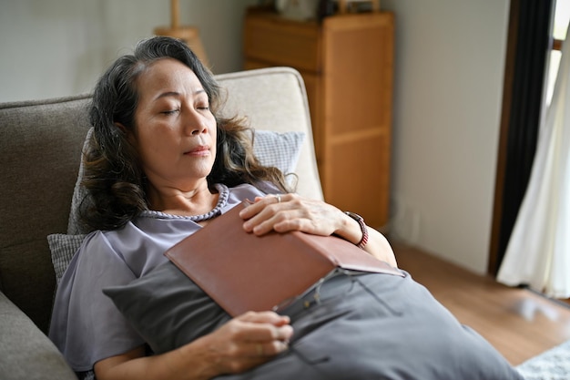 Una anciana asiática tranquila durmiendo en un sofá en su sala de estar se queda dormida mientras lee un libro