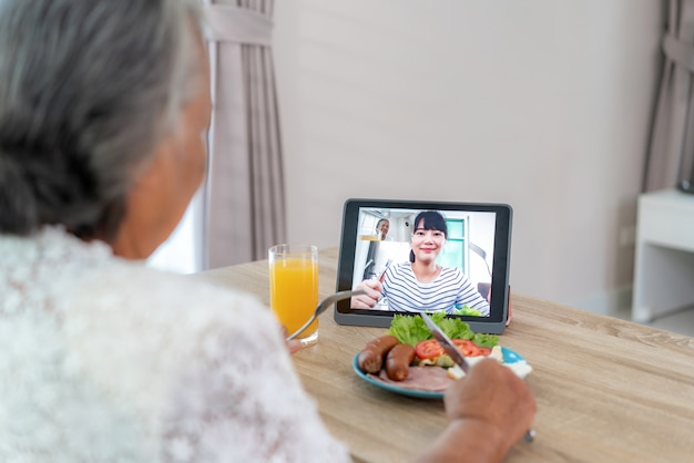 Anciana asiática virtual happy hour reunión y comer alimentos en línea junto con su hija en video conferencia con tableta digital para una reunión en línea en video llamada