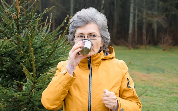 Anciana bebiendo té caliente en el bosque al aire libre. Retrato de una mujer mayor caminando en clima frío.