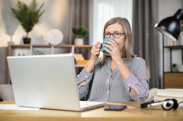 Anciana bebiendo té mientras estudia el informe en la computadora portátil