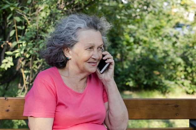 Una anciana con cabello gris está hablando por teléfono mientras está sentada en un banco. Abuela con reliquias y pecas sonríe mientras habla por teléfono.