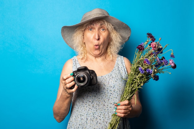 Anciana con cara de sorpresa en un vestido y sombrero de ala ancha sosteniendo un ramo de flores y una cámara sobre una superficie azul