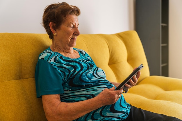 Foto anciana caucásica sentada en un sofá amarillo leyendo con un libro electrónico lleva una camiseta azul