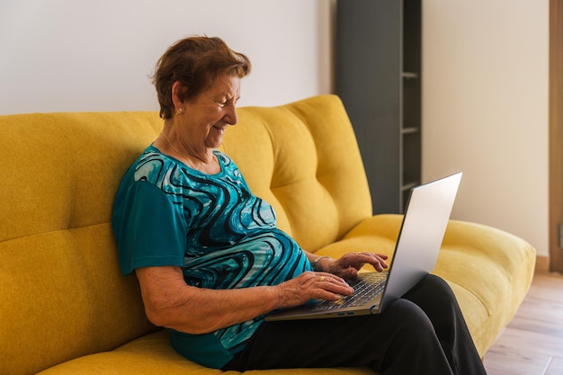 Una anciana caucásica sonriendo está escribiendo en una laptop sentada en un sofá amarillo
