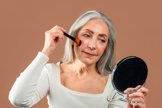 Una anciana caucásica sonriente con el pelo gris aplicando rubor en las mejillas mira en el espejo aislado en el fondo marrón