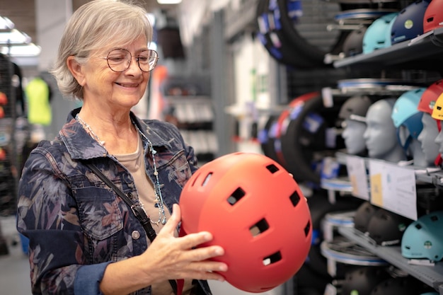 Anciana en una chaqueta de mezclilla selecciona un casco protector de bicicleta en una tienda de deportes