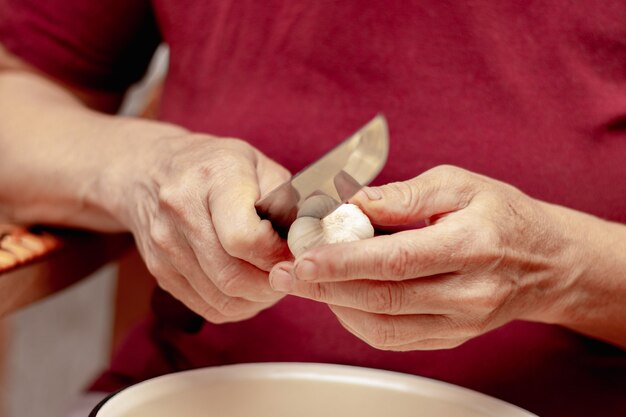 Anciana en la cocina corta el ajo con un cuchillo de cocina. Trabajar en la cocina, cocinar