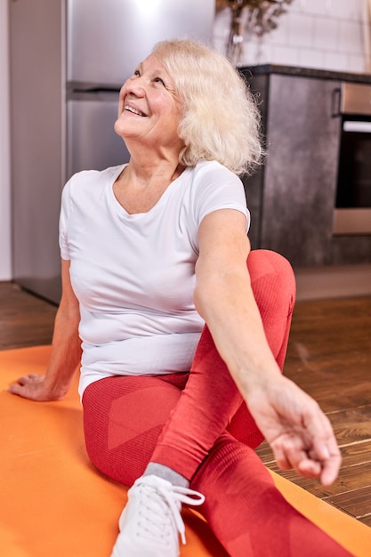 Anciana disfruta de ejercicios deportivos en el suelo en casa, se sienta con las piernas cruzadas y mirando hacia arriba, sonriendo