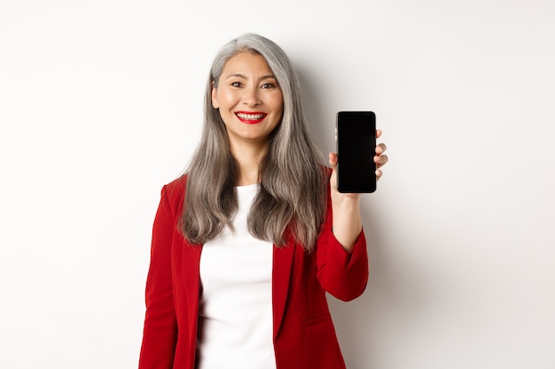 Anciana empresaria en chaqueta roja, mostrando la pantalla del teléfono inteligente en blanco y sonriendo, demostrar la aplicación móvil, fondo blanco.