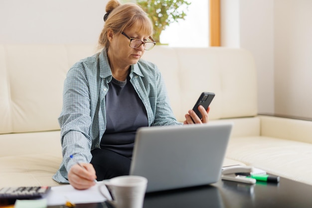 Anciana empresaria que trabaja desde la oficina en casa hablando por teléfono móvil riéndose disfrutando de una conversación telefónica usando una aplicación en línea en una computadora portátil para consultar al cliente