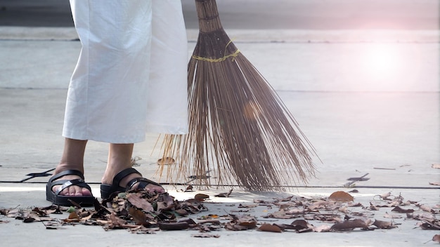 La anciana está barriendo hojas secas en el piso de cemento al aire libre con una gran escoba larga.