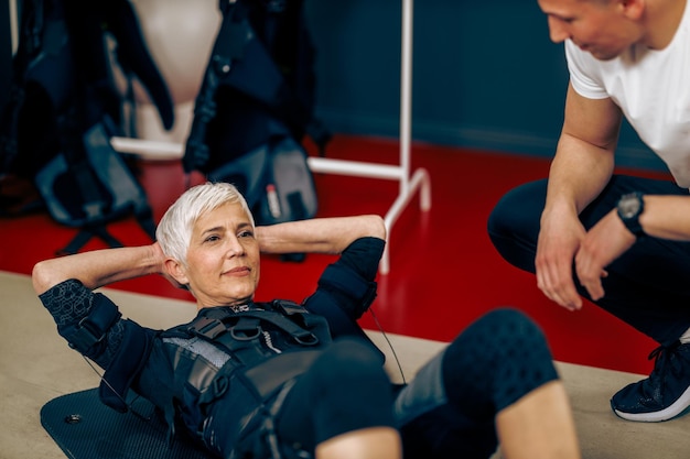 La anciana está haciendo ejercicios de abdominales durante el entrenamiento EMS con entrenador personal en el gimnasio.