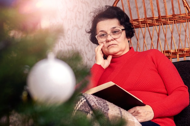 Una anciana está leyendo cuidadosamente la Biblia cerca del árbol de Navidad Celebrando la Navidad sola