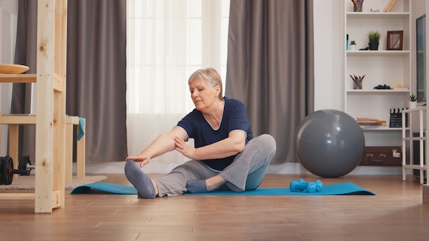 Anciana estirando las piernas sentado en la estera de yoga en la sala de estar. Activo estilo de vida saludable deportivo anciano entrenamiento entrenamiento bienestar en el hogar y ejercicio en interiores