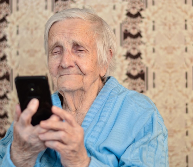 Una anciana feliz de 90 años con gafas y una chaqueta azul sonríe usando un teléfono inteligente