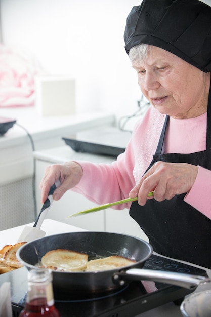 Una anciana con gorra y delantal friendo panqueques en la cocina