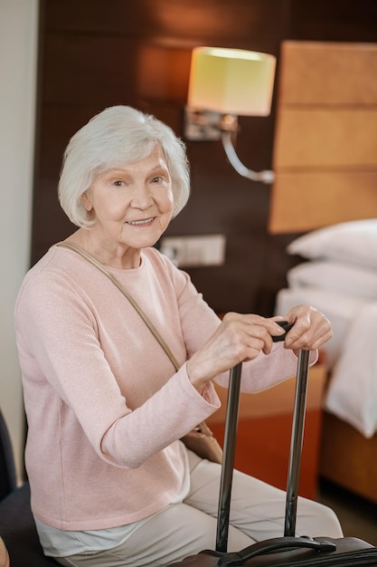 Una anciana guapa sentada con una maleta en una habitación de hotel