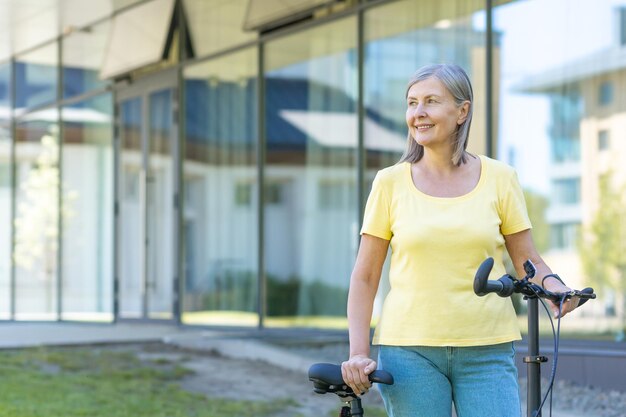 Una anciana jubilada activa camina fuera de un edificio moderno con una bicicleta