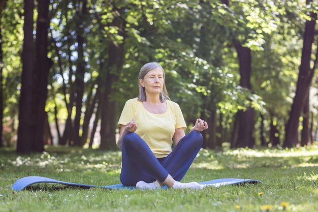 Una anciana jubilada de pelo gris en el parque medita y realiza ejercicios de respiración el día de verano en posición de loto