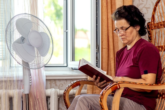 Una anciana lee la Biblia sentada en una silla junto a una ventana abierta y un ventilador