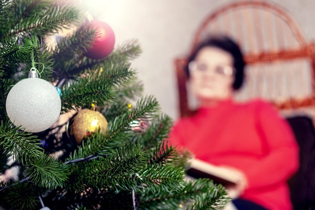 Una anciana lee un libro, la Biblia en una silla de mimbre frente a un árbol de Navidad Celebrando la Navidad sola