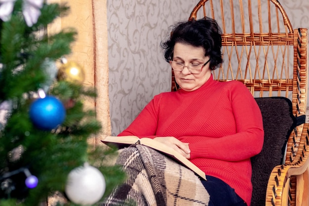 Una anciana lee un libro, la Biblia en una silla de mimbre frente a un árbol de Navidad Celebrando la Navidad sola