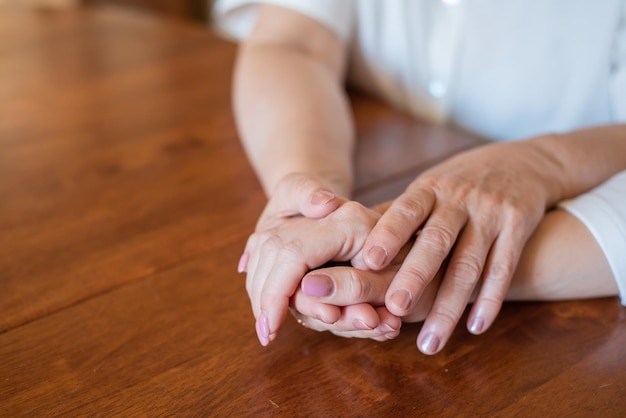 La anciana madre y su hija se toman de la mano mientras están sentadas en la mesaCerca de mujeres de diferentes generaciones tomándose de la mano Primer plano de las manos de madre e hija sosteniendo