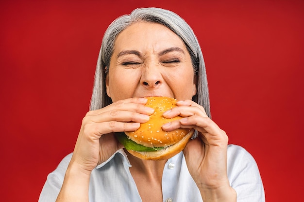 Una anciana madura comiendo hamburguesas con satisfacción La abuela disfruta de una sabrosa hamburguesa para llevar delicioso bocado de hamburguesa para pedir comida rápida mientras está hambrienta aislada sobre un fondo rojo