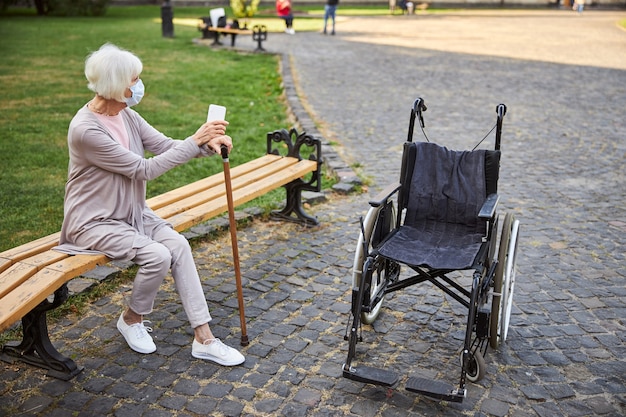 Anciana con máscara médica sentada en un banco con las manos en un bastón y mirando a otro lado. Silla de ruedas vacía frente a ella