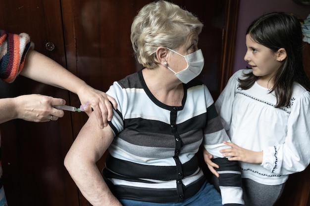 Anciana con mascarilla quirúrgica recibiendo la vacuna Covid19 y nieta a su lado apoyándola