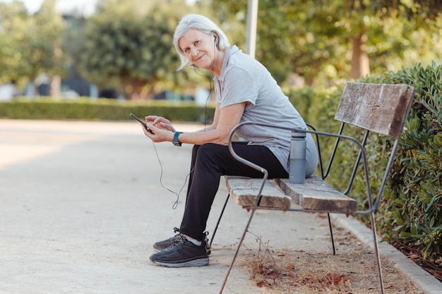 Una anciana mira una cámara sonriente mientras sostiene un teléfono móvil sentado en un banco del parque