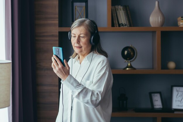 La anciana moderna en casa junto a la ventana escucha música en el teléfono usa una aplicación de música para escuchar en línea y auriculares