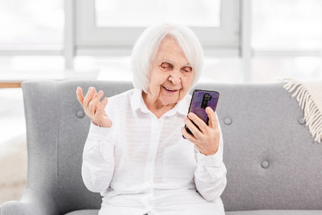 Anciana moderna sentada en el sofá en casa y hablando con amigos por video con smartphone