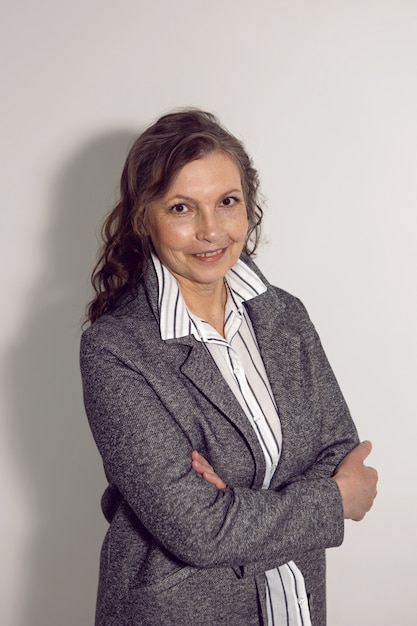 Anciana mujer de negocios con una camisa blanca y una chaqueta gris está contra una pared blanca