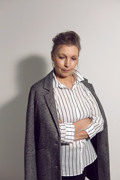 Anciana mujer de negocios con una camisa blanca y una chaqueta gris está contra una pared blanca