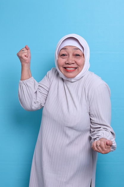 Una anciana musulmana asiática de los años 60 celebra apretar los puños aislada de fondo azul