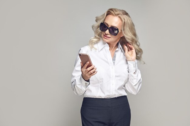 Una anciana de negocios con un aspecto elegante y estricto y gafas de sol se para sobre gris y sostiene el teléfono