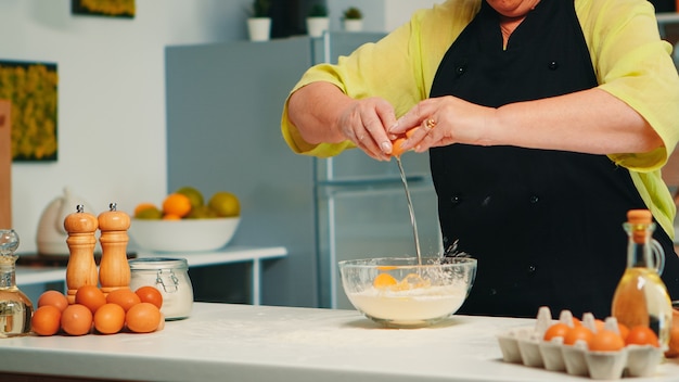 Anciana panadera romper huevos en un tazón de vidrio para una receta de comida sabrosa en la cocina de casa. chef anciano jubilado con bonete mezclando a mano, amasando ingredientes de pastelería para hornear pasteles caseros.