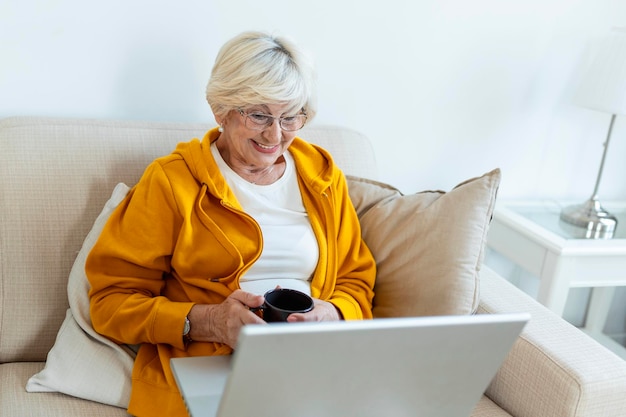 Una anciana pasa el tiempo libre en casa con una computadora portátil Una anciana sonriente se sienta en un sofá acogedor con una taza de café o té Navegación web viendo series de televisión de compras en una videollamada en red
