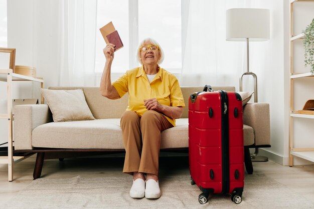 Anciana con pasaporte y boleto de viaje empacó una maleta roja de vacaciones y atención médica Sonriente anciana felizmente sentada en el sofá antes del viaje levantó las manos para la alegría de la jubilación