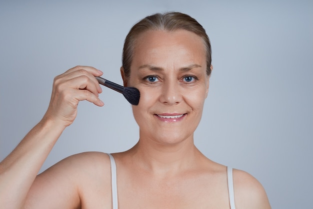 Una anciana se pone cosméticos decorativos en las mejillas. Cuidado de la piel de la cara en la vejez.