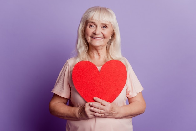 Anciana positiva mantenga gran tarjeta de corazón de San Valentín aislada sobre fondo violeta