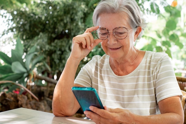 Una anciana relajada sentada en un jardín al aire libre usando un teléfono móvil anciana con anteojos sonriendo leyendo un mensaje en un smartphone