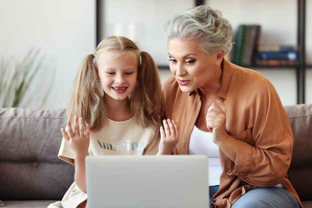 Anciana sacudiendo el dedo y explicando datos a una niña sonriente mientras se sienta en el sofá y ve un curso en línea en una computadora portátil en casa