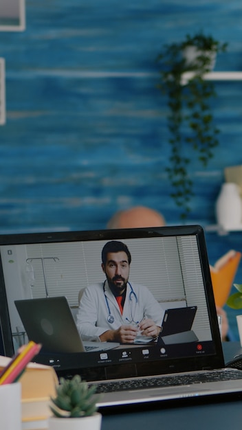 Foto anciana sentada a la mesa en casa mirando la computadora portátil respondiendo preguntas sobre conversaciones de salud con el terapeuta mediante la aplicación de llamada de videoconferencia, sosteniendo el matraz de píldoras concepto de consulta médica remota